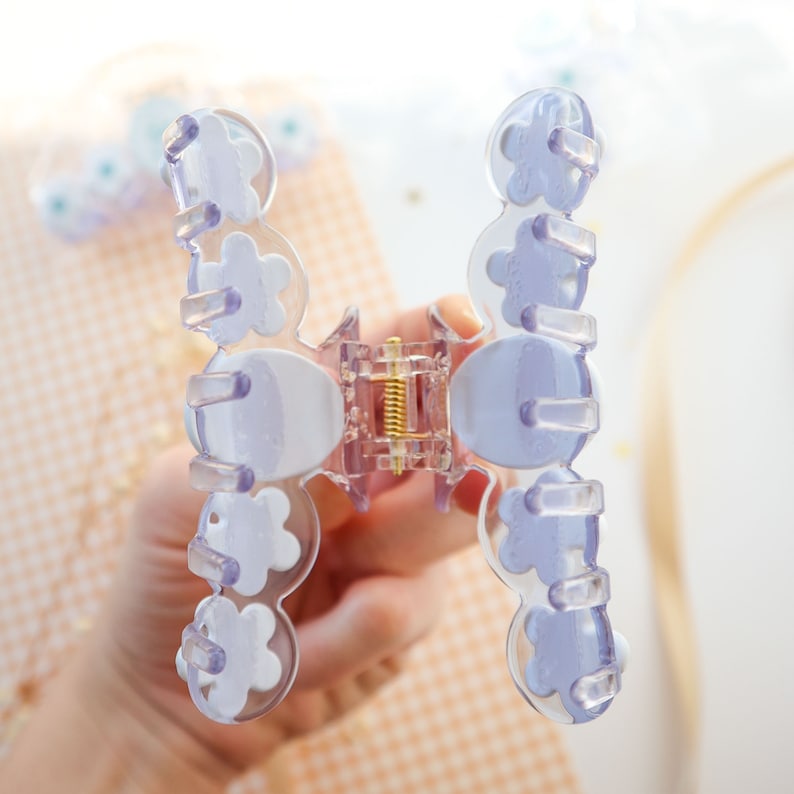 May include: Clear plastic hair claw clip with a light purple and white floral design. The clip has a gold-colored spring mechanism and is designed to hold hair in place. A stylish hair accessory.