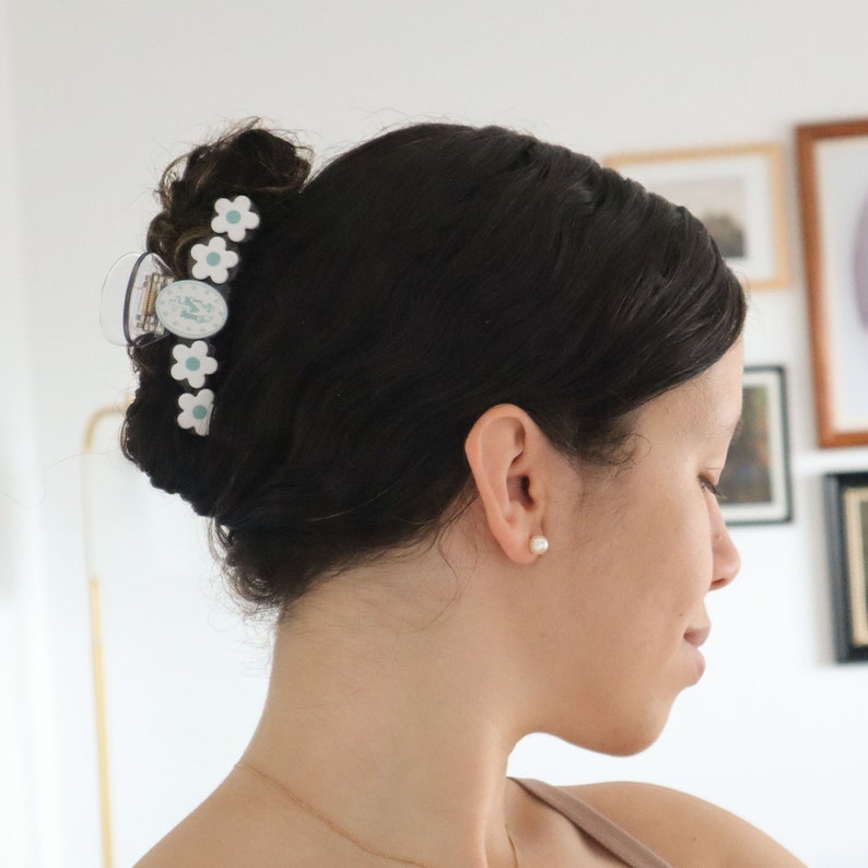 May include: A clear plastic hair claw clip with white daisy-shaped accents and a central oval design. The clip is holding up dark hair in a bun. A woman is wearing a pearl earring and a gold necklace.