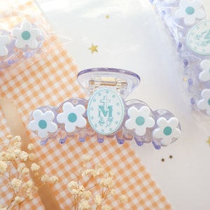 May include: A clear, plastic hair claw clip with white flower accents and a central oval emblem. The emblem features a cross, the letter "M", and other symbols in light blue. The clip is designed to hold hair in place.