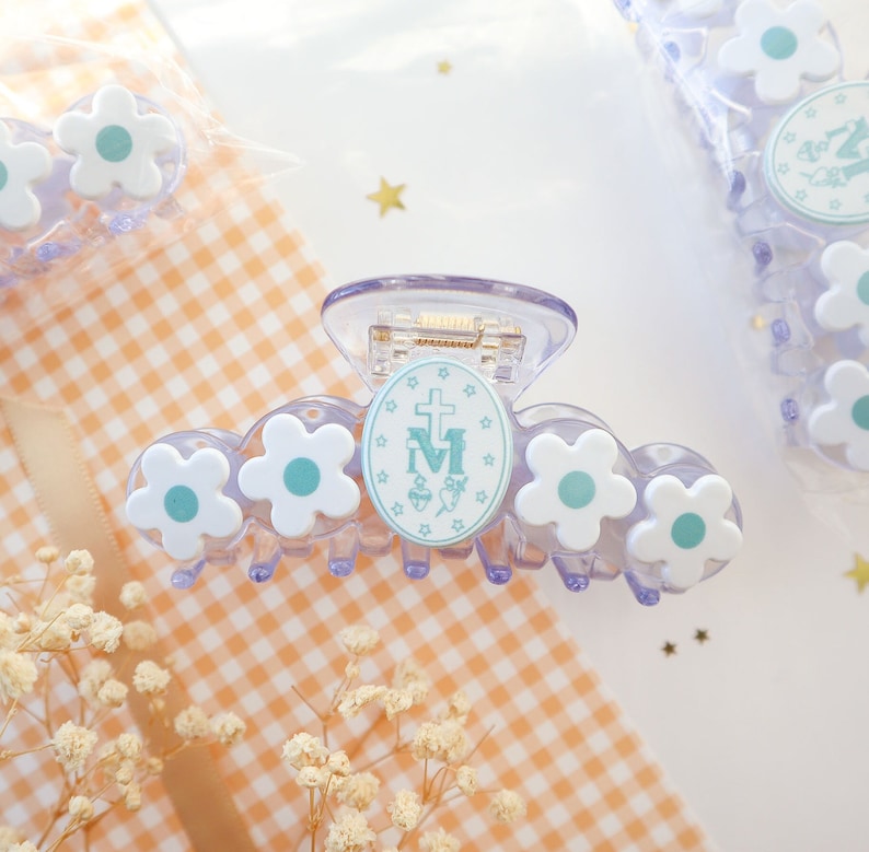 May include: A clear, plastic hair claw clip with white flower accents and a central oval emblem. The emblem features a cross, the letter "M", and other symbols in light blue. The clip is designed to hold hair in place.