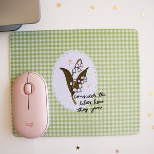 May include: A pink wireless Logitech mouse rests beside a green gingham mousepad with a lily of the valley illustration and the text "consider the lilies, how they grow." Gold star confetti decorates the white surface.