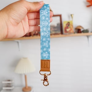 Pode incluir: Um cordão azul claro com um padrão branco e uma alça de couro marrom. O cordão tem um clipe de metal dourado.