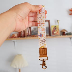 Puede incluir: Un cordón blanco con un patrón floral y un cierre de metal dorado. El cordón tiene una correa de cuero marrón en la parte superior.