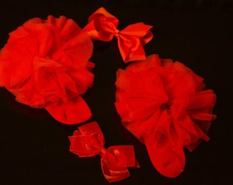 May include: A pair of bright red socks featuring large, voluminous tulle ruffles at the cuffs, accompanied by two matching red satin hair bows. The set is presented against a dark background, highlighting the vibrant red color and texture.
