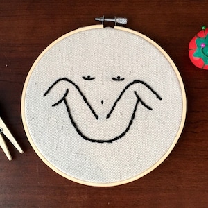 May include: An embroidery hoop with a beige fabric featuring a minimalist black stitched face. A wooden clothespin holds black thread, and a tomato-shaped pin cushion with colorful pins sits nearby. The wooden hoop is approximately 6 inches in diameter.