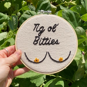 May include: A white embroidery hoop with the text "'Tis ol' Bitties" embroidered in black thread. Two embroidered breasts are depicted with yellow flower accents.