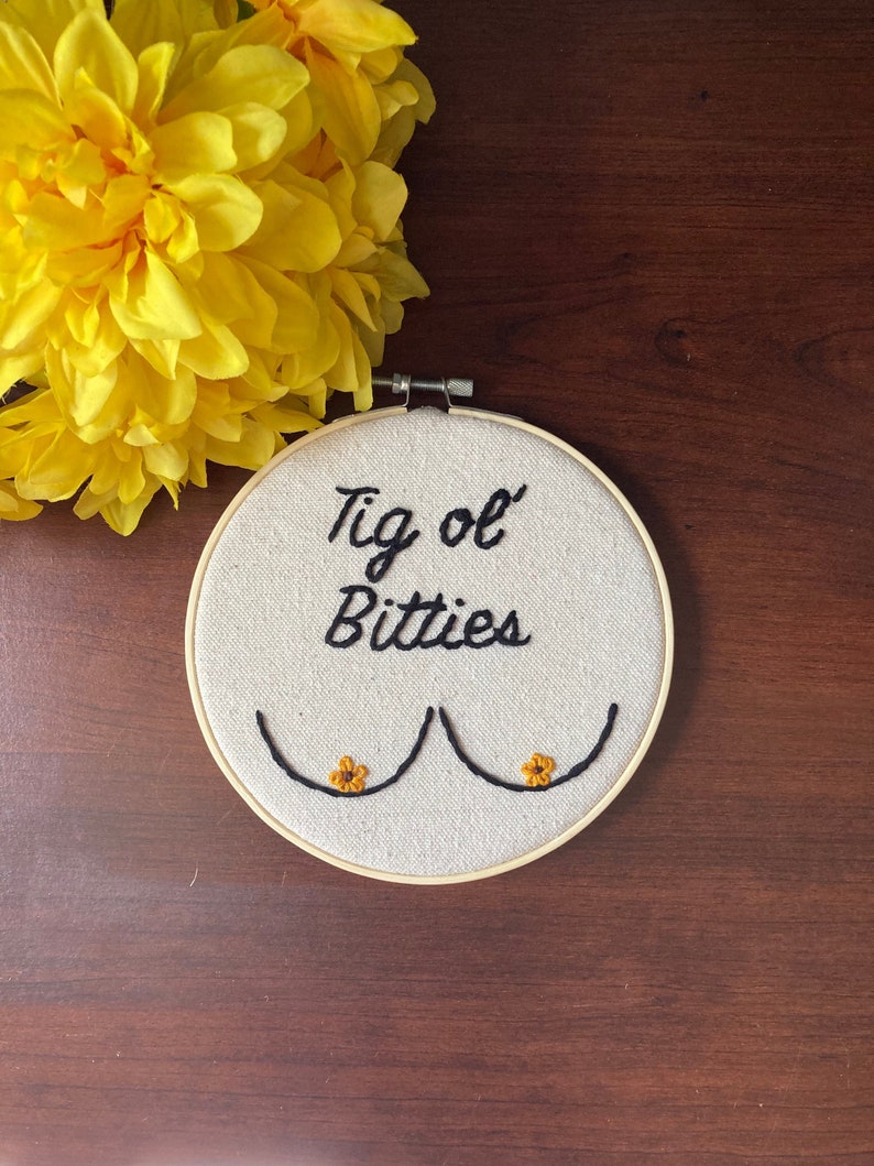 May include: A white embroidery hoop with the phrase "Tig ol' Bitties" embroidered in black thread. Two small yellow embroidered flowers are on the bottom of the hoop.