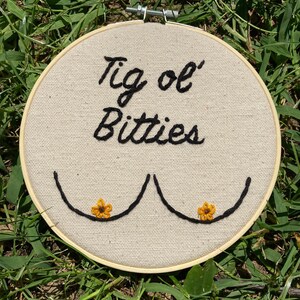 May include: Embroidered hoop art with the text "Tig ol' Bitties" and two stylized breasts with yellow flowers.