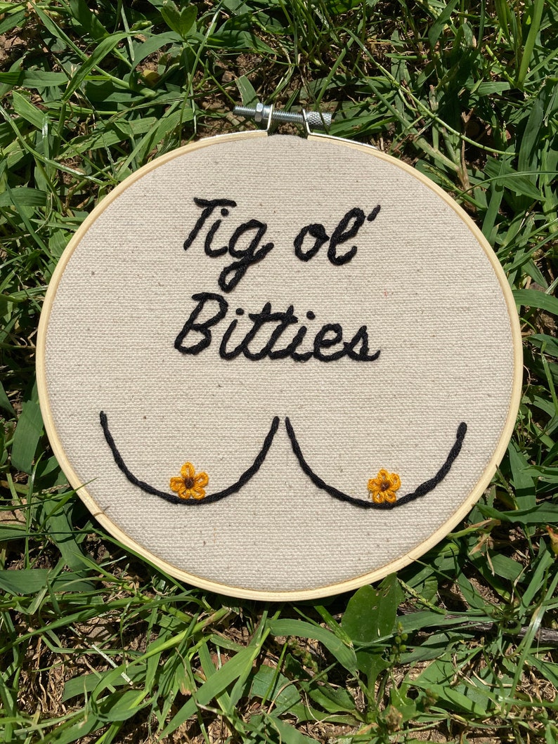 May include: Embroidered hoop art with the text "Tig ol' Bitties" and two stylized breasts with yellow flowers.
