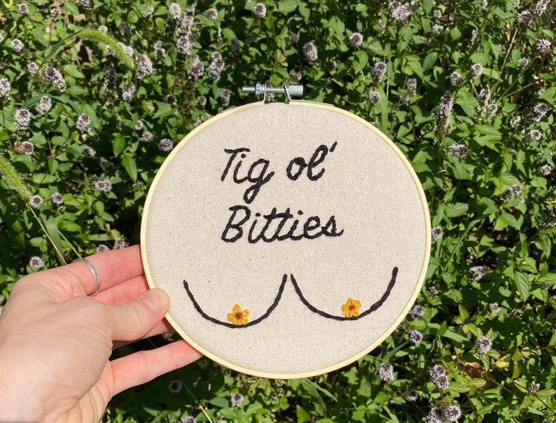 May include: A white embroidery hoop with the text "Tig ol' Bitties" embroidered in black thread. Two small yellow embroidered flowers are on either side of the text.