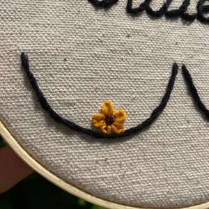 May include: Embroidered hoop art with the word "Bittie" in black thread and a small yellow flower with a brown center.