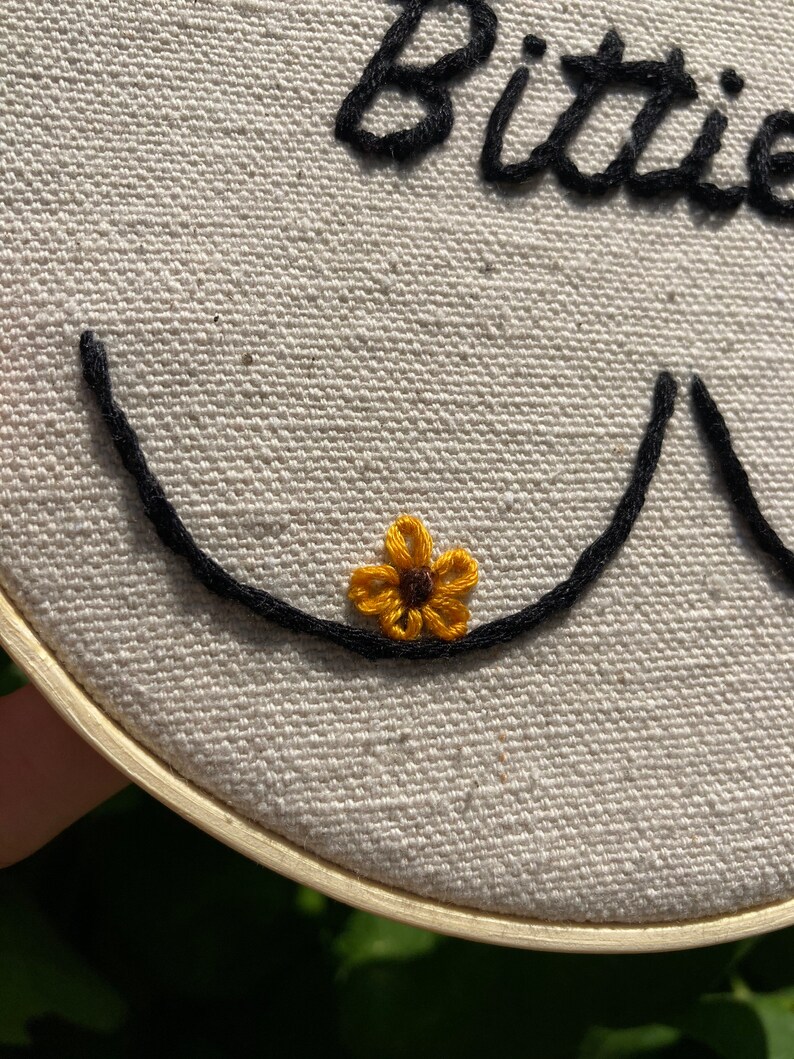 May include: Embroidered hoop art with the word "Bittie" in black thread and a small yellow flower with a brown center.