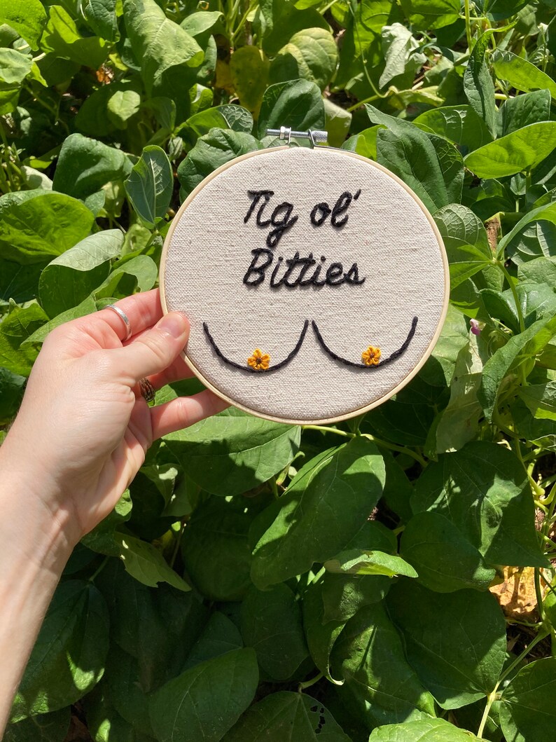May include: A white embroidery hoop with the text "'Tis ol' Bitties" embroidered in black thread. Two embroidered breasts are depicted with yellow flower accents.
