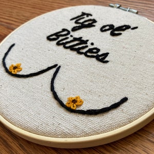 May include: Embroidered hoop art with the text "Big ol' Bitties" in black cursive script. Two yellow embroidered flowers are on the embroidered breasts.