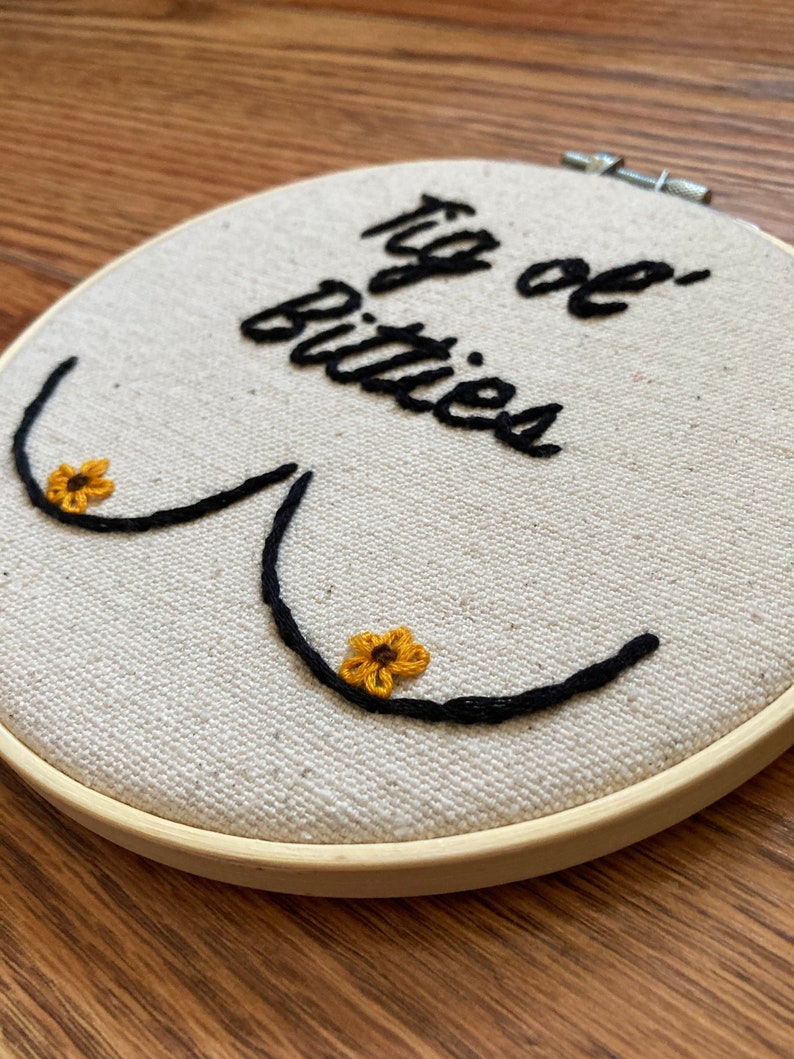 May include: Embroidered hoop art with the text "Big ol' Bitties" in black cursive script. Two yellow embroidered flowers are on the embroidered breasts.