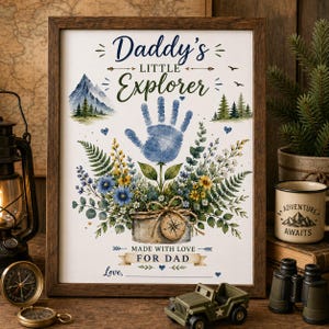 May include: A framed print with the text "Daddy's Little Explorer" and a blue handprint design. The artwork includes watercolor illustrations of mountains, flowers, and a compass, with the message "Made with Love For Dad."