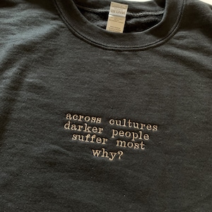 Op de afbeelding: Zwart sweatshirt met de tekst "across cultures darker people suffer most why?" geborduurd in gouden draad.