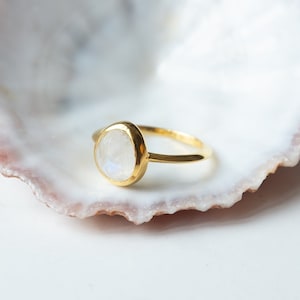 Könnte beinhalten: Ein goldener Ring mit einem ovalen weißen Mondstein, der in eine schlichte Bandfassung gefasst ist. Der Ring liegt auf einer weißen Muschel.