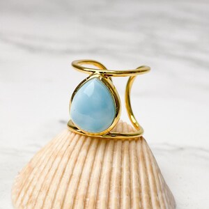 Könnte beinhalten: Ein goldener Ring mit einem hellblauen, tropfenförmigen Edelstein. Der Ring wird auf einer Muschel präsentiert.