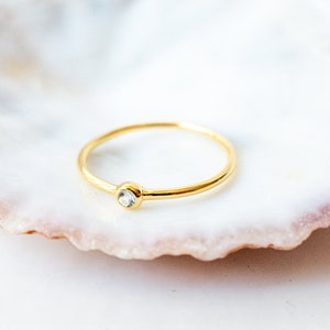 Puede incluir: Un anillo de oro simple con una sola piedra transparente engastada en la banda. El anillo está apoyado en una concha blanca.