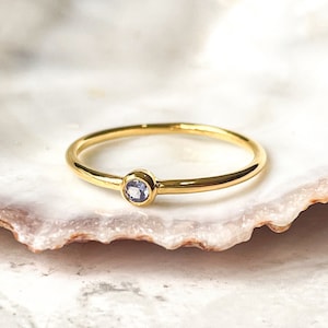 Può includere: Un delicato anello d'oro con una piccola pietra preziosa di colore viola chiaro incastonata al centro.