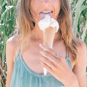 May include: A woman with long brown hair is eating a white ice cream cone. She is wearing a light blue tank top and has a gold necklace on.
