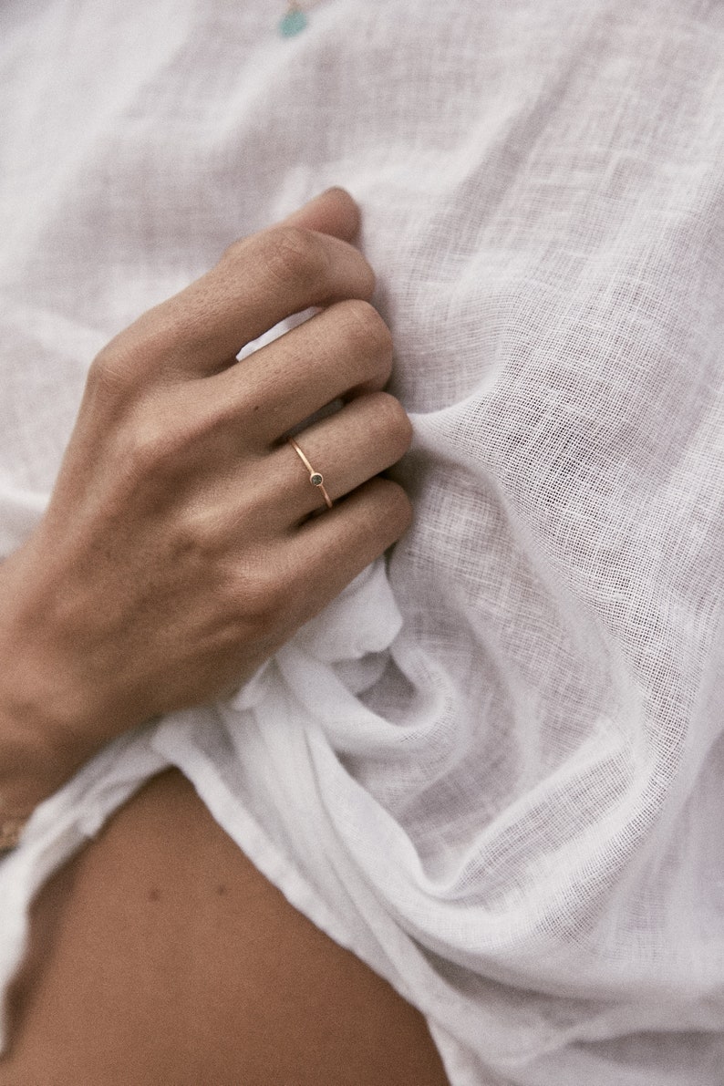 May include: A delicate gold ring with a small black gemstone. The ring is worn on the finger of a person's hand.