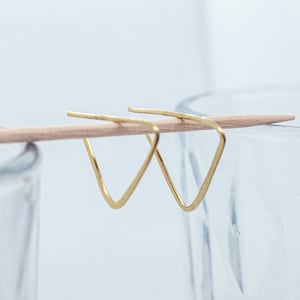 Può includere: Un paio di orecchini a forma di triangolo dorati. Gli orecchini sono semplici e minimalisti.