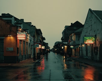 Può includere: Una stretta strada bagnata a New Orleans, Louisiana, con edifici in mattoni su entrambi i lati. La strada è fiancheggiata da insegne al neon, tra cui "Slush" e "Tropical Isle".