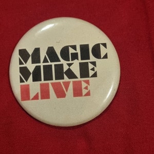 Op de afbeelding: Een witte knop met zwarte en rode letters met de tekst "MAGIC MIKE LIVE".