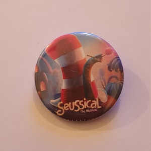 May include: A round button with a colorful image of a character from the musical Seussical. The character is wearing a red and white striped hat and is holding a flower. The text "Seussical The Musical" is printed on the button.
