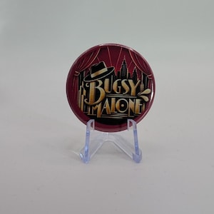Può includere: Una spilla rotonda con sfondo rosso scuro, caratterizzata da un grafico con un cappello fedora nero, la sagoma di uno skyline cittadino e il testo color oro che recita Bugsy Malone, esposta su un supporto in plastica trasparente.