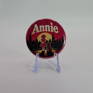 Peut inclure: Un badge rond épinglé portant le mot Annie en lettres blanches. L'illustration représente la silhouette d'une personne et d'un chien devant un soleil jaune et une ligne d'horizon urbaine sombre sur fond rouge foncé, posé sur un support transparent.
