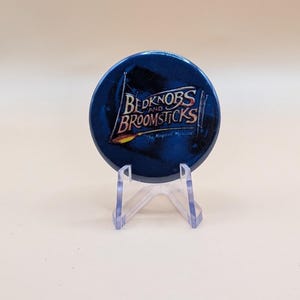 May include: A circular blue pinback button featuring the text Bedknobs and Broomsticks The Magical Musical in white and gold lettering. The design includes a graphic of a broomstick and a flag. The button is displayed on a clear plastic stand.