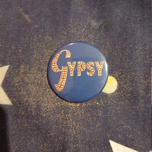 Peut inclure: Un bouton bleu avec un logo "Gypsy" blanc et rouge avec des points dorés autour des lettres.