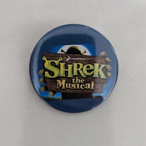 Könnte beinhalten: Ein blauer Knopf mit einem Bild von Shrek dem Musical. Das Logo befindet sich auf einem Holzschild mit den Worten "Shrek das Musical" in gelben Buchstaben.