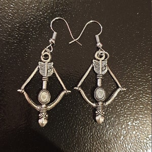 May include: A pair of silver bow and arrow earrings. The earrings are made of metal and feature a detailed design. The bow and arrow are in a classic style.