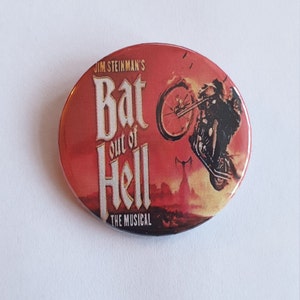 May include: A red, white and black button featuring a motorcycle jumping over a fiery cityscape. The text "Jim Steinman's Bat Out of Hell The Musical" is printed on the button.