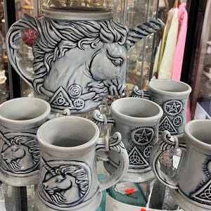 May include: A ceramic tea set featuring a large unicorn-shaped teapot and four matching mugs. The pieces are light gray with dark blue detailing, showcasing embossed unicorn heads and geometric symbols including pentagrams and triquetra knots.