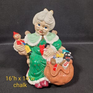 May include: A chalk-painted figurine of a seated woman wearing glasses and a green dress, holding dolls and a sack of toys. The figurine is 40.6 cm high and 35.6 cm wide. The text "16"h x 14"w chalk" is visible.