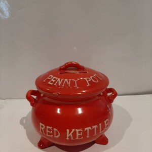 May include: Red ceramic piggy bank shaped like a kettle with a lid. The lid has a slot for coins and the words "Penny Pot" written on it. The body of the piggy bank has the words "Red Kettle" written on it.