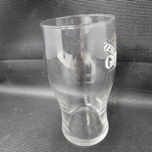 May include: A clear, tulip-shaped pint glass with the Guinness logo in white. The glass has a curved base and a slightly flared top. The glass is empty and appears to be clean.