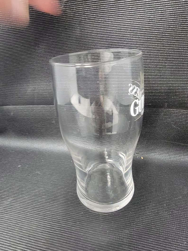 May include: A clear, tulip-shaped pint glass with the Guinness logo in white. The glass has a curved base and a slightly flared top. The glass is empty and appears to be clean.