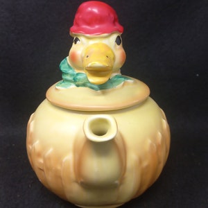 Könnte beinhalten: Eine gelbe Teekanne aus Keramik mit einem Entenkopfdeckel. Die Ente trägt einen roten Hut, einen grünen Schal und einen gelben Schnabel. Die Teekanne hat einen braunen Henkel und einen braunen Ausguss.