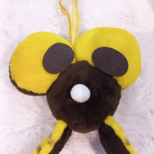 Może przedstawiać: Pluszowa zabawka w kolorze brązowym i żółtym z dużymi, okrągłymi, czarnymi oczami i białym nosem. Zabawka ma dwa długie, zwisające uszy i żółtą sznurówkę przymocowaną do górnej części głowy.