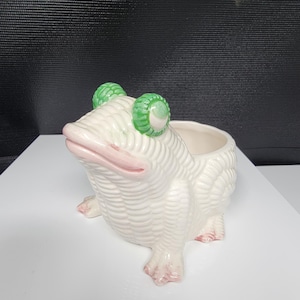 Puede incluir: Jardinera de cerámica blanca con forma de rana, con ojos verdes y detalles rosas. La jardinera tiene un diseño texturizado y acanalado y una amplia abertura para plantas. La rana está sentada, con sus patas delanteras visibles.