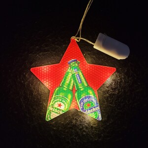 May include: A red star-shaped light featuring two green Heineken beer bottle images. One bottle says "Heineken Original" and the other says "Bi&egrave;re Sans Alcool". The light is powered by a small battery pack.
