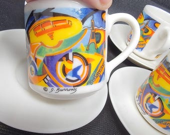 May include: A set of four small white ceramic espresso cups and saucers. The cups have a colourful abstract design with yellow, blue, and green elements, including an image of the Eiffel Tower. The artist's signature, "D. Burrows," is visible.