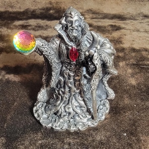 May include: A detailed silver-colored figurine of a wizard, holding a colorful orb in one hand and a staff in the other. The wizard wears a long robe and has a long beard. A red gem is set into the wizard's chest. The figurine is a fantasy-themed collectible.