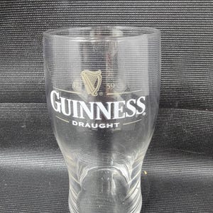 May include: A clear pint glass with the Guinness logo, featuring a harp and the word "Guinness" in white lettering. The word "Draught" is printed below. The glass has a slightly curved shape and a wide mouth.